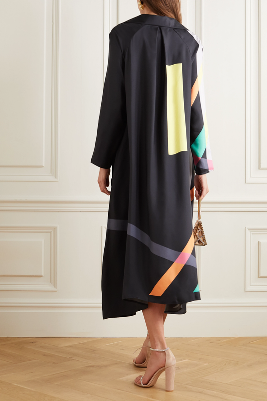 Woman wearing a long black coat with colorful geometric patterns, standing in a room with white paneled walls.