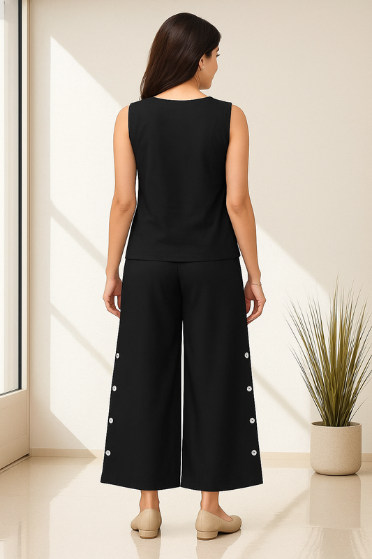 Woman wearing a black sleeveless top and wide-leg pants with white buttons in a bright room.