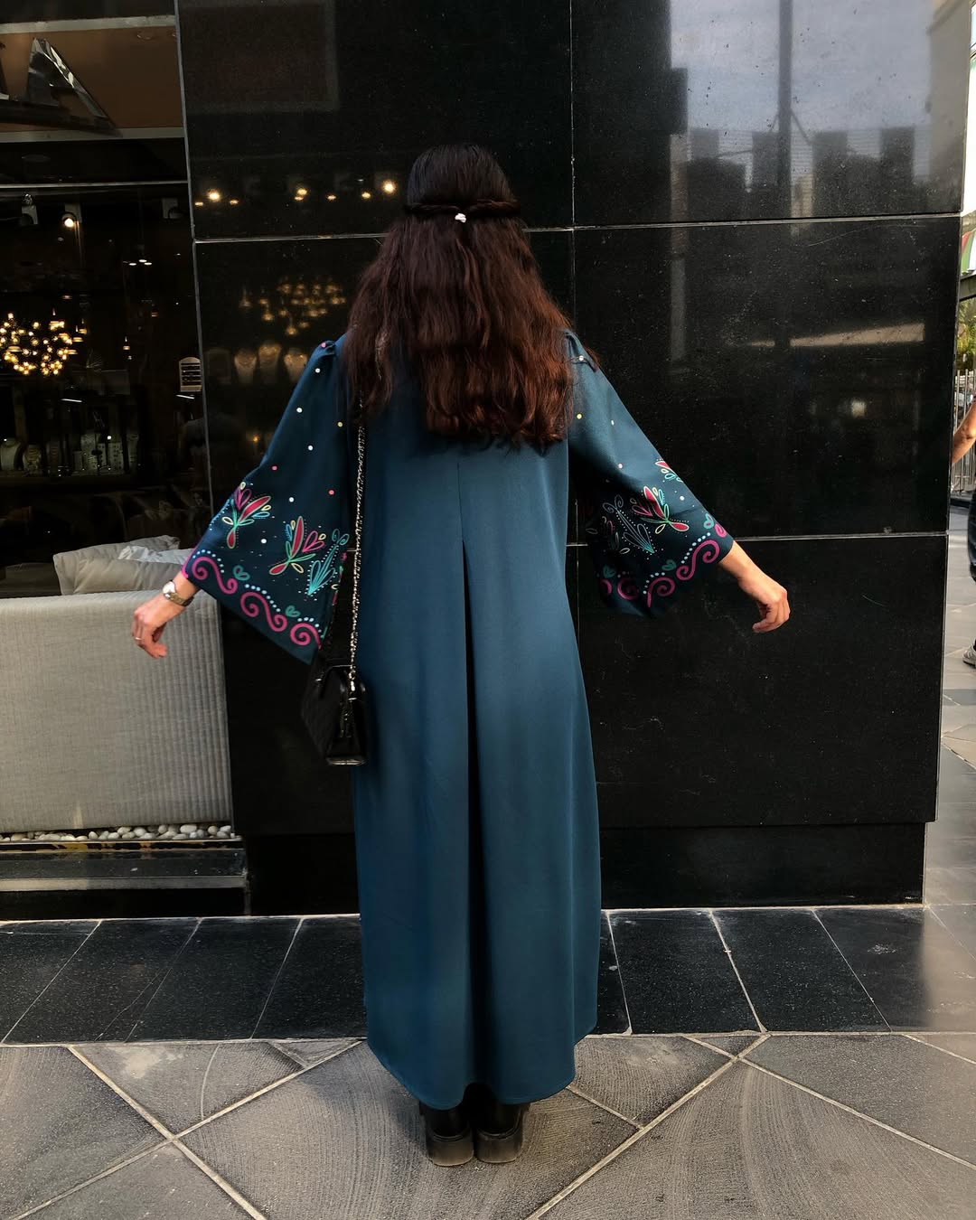 A woman viewed from behind wearing a long teal blue dress with wide embroidered sleeves, standing outdoors in front of a black reflective building facade.