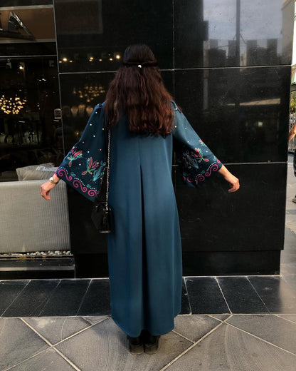 A woman viewed from behind wearing a long teal blue dress with wide embroidered sleeves, standing outdoors in front of a black reflective building facade.