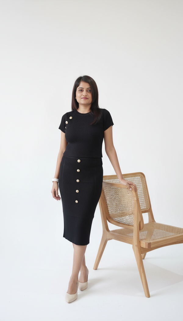 Woman wearing black ribbed co-ord set with pearl button detailing, featuring a fitted crop top and midi pencil skirt, styled with nude flats against a brown background.